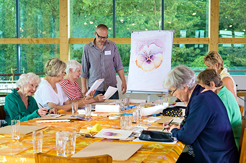 Group of six female senior citizens at long table in painting class with male teacher in glasses standing near front.